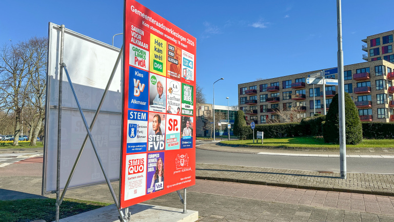 Verkiezingsbord met posters van politieke partijen voor de gemeenteraadsverkiezingen 2026 in Alkmaar, langs een straat.