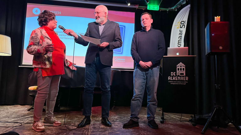 Drie personen op een podium, waarvan één een microfoon vasthoudt en een vrouw aanspreekt; achtergrond met projectiescherm.