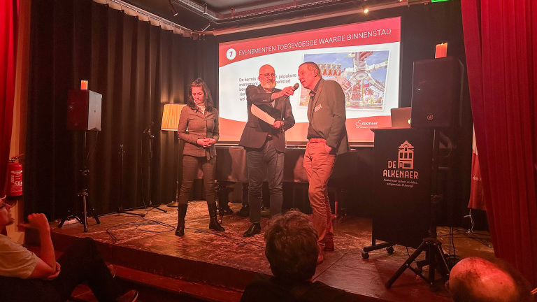 Drie mensen staan op een podium met een presentatie over evenementen in de binnenstad, in een zaal met publiek.