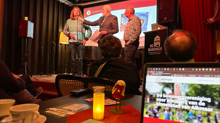 Drie mannen op een podium tijdens een presentatie in een zaal met publiek, een scherm en een laptop met tekst over Alkmaar.