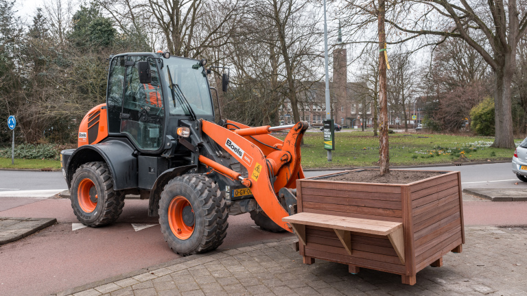 Graafmachine van Boels plaatst een houten plantenbak met boom op een trottoir nabij een rotonde.