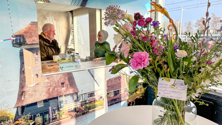 Een boeket kleurrijke bloemen in een vaas, met op de achtergrond twee mensen die achter een toonbank staan.