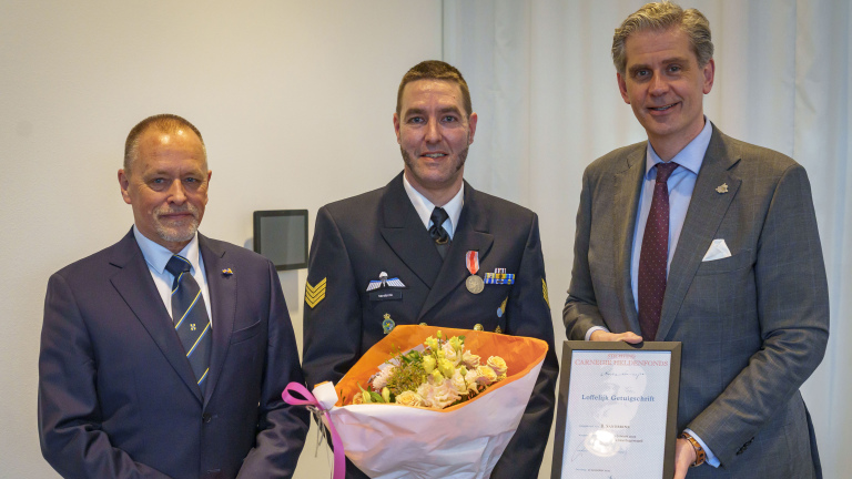 Drie mannen in formele kleding; de middelste man heeft een medaille en bloemen, de man rechts houdt een certificaat.
