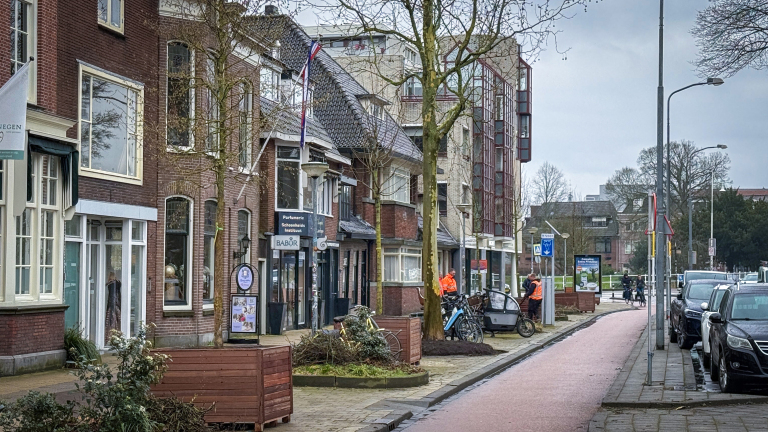 Straatbeeld met winkels, een paar mensen in oranje werkkleding, fietsen, en geparkeerde auto's aan de rechterkant.