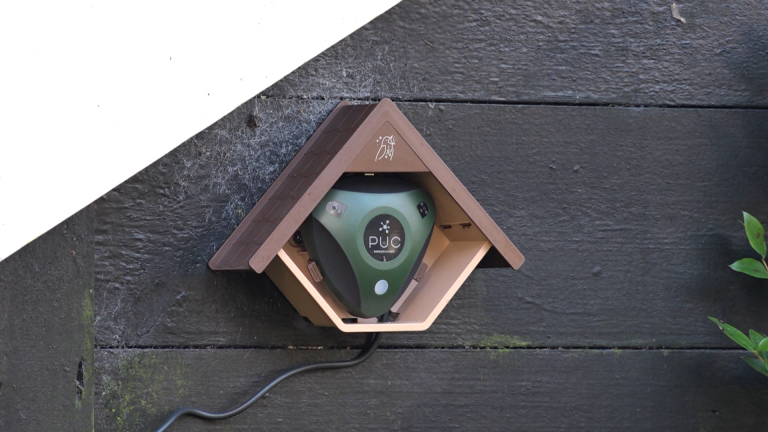 Voederhuisje met een groen apparaat erin, gemonteerd op een donkere, houten muur. Er zit een stroomkabel aan vast.