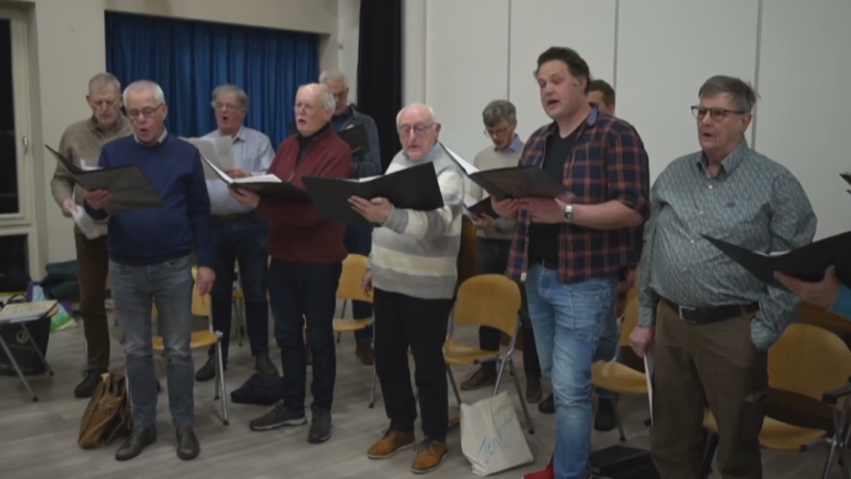 Mannenenkoor aan het repeteren in een zaal, met zeven staande mannen die muziekpartituren vasthouden.