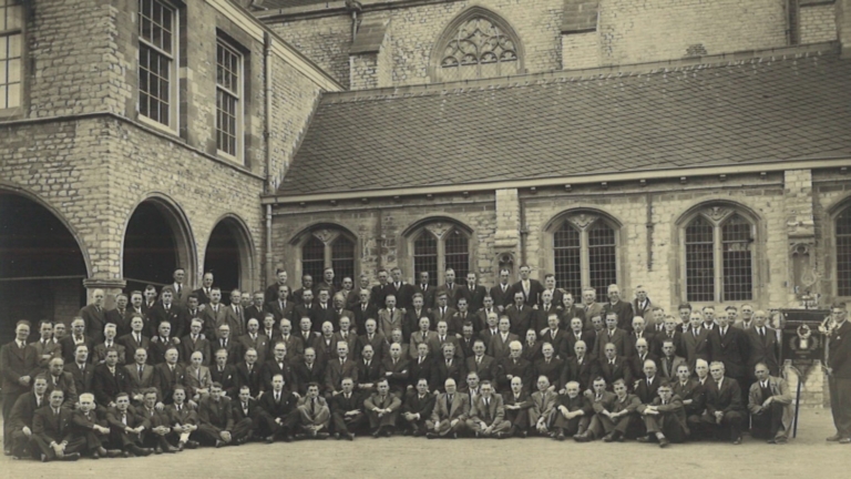Een grote groep mannen poseert voor een historisch gebouw met ramen in gotische stijl.