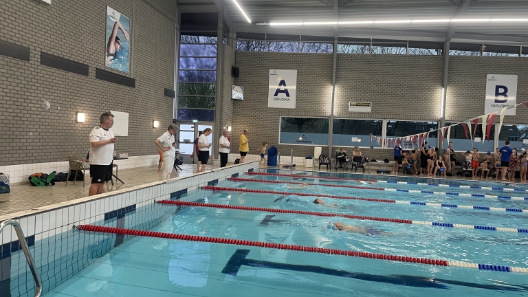 Zwemwedstrijd in een overdekt zwembad met juryleden langs de kant en toeschouwers op de achtergrond.