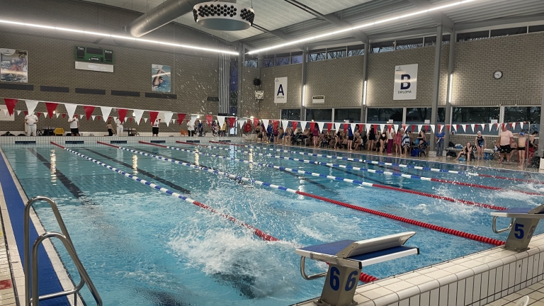 Binnenzwembad met meerdere zwembanen, waar mensen op de startblokken staan en anderen aan de kant toekijken.