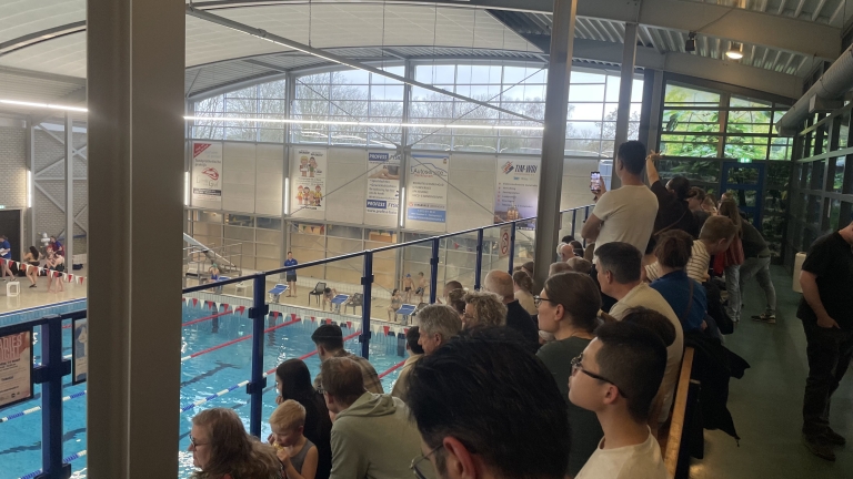 Toeschouwers zitten op tribunes bij een zwembad, waar een zwemwedstrijd lijkt te plaatsvinden.