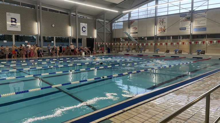 Zwembad met meerdere zwemmers in banen; toeschouwers staan langs de kant onder borden met letters "B" en "C".