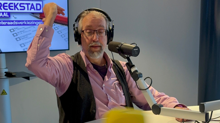 Man in studio met koptelefoon voor microfoon, maakt een gebaar met hand. Achtergrond bevat tekst over verkiezingen.