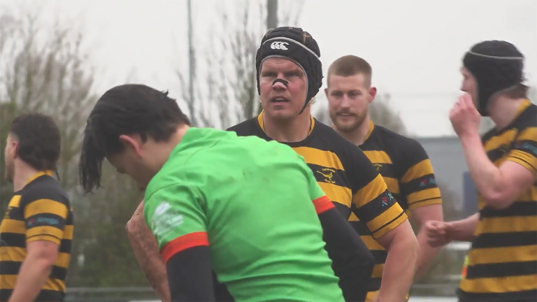 Een rugbyspeler met een zwart-geel gestreept shirt en neustape staat op het veld, omringd door medespelers tijdens een wedstrijd.