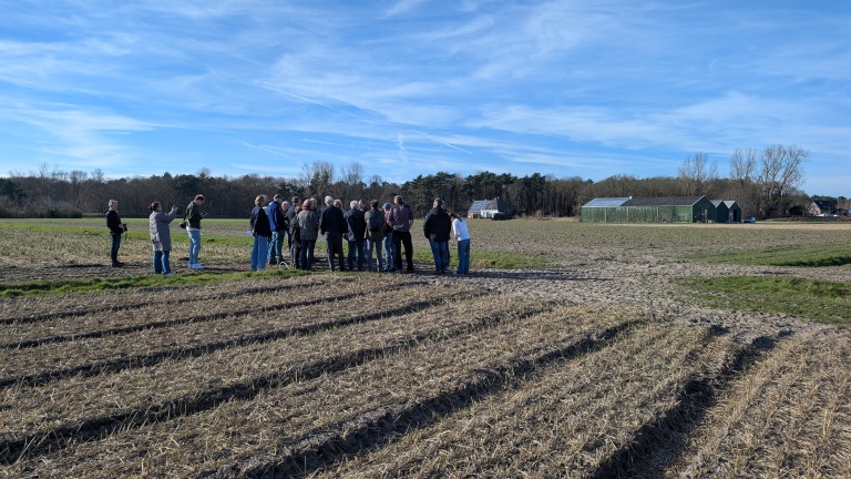 Een groep mensen staat op een akker bij een kas en een boerderij, onder een blauwe lucht.
