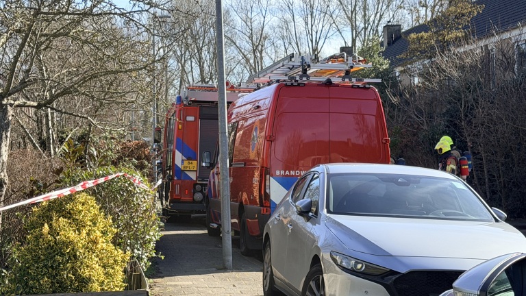 Brandweerwagens en een brandweerman in een woonwijk, met afzetlint en geparkeerde auto's op de voorgrond.