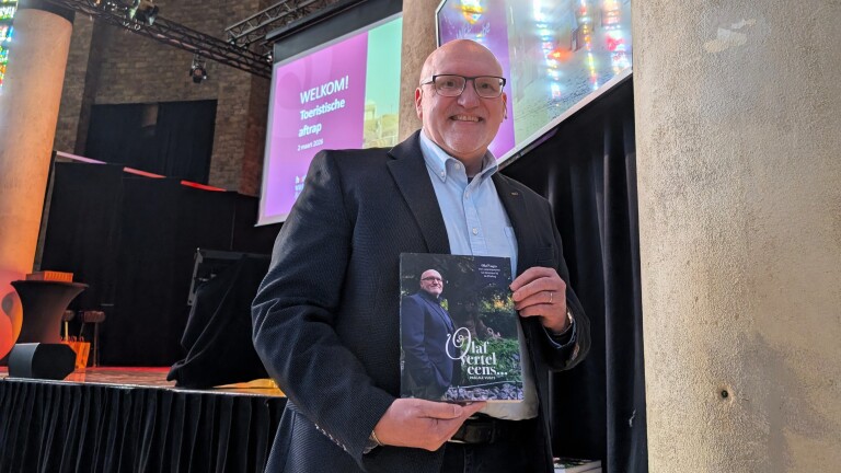 Man in een donkere blazer houdt een boek vast; op de achtergrond een projector met de tekst "Welkom! Toeristische aftrap, 2 maart 2026".