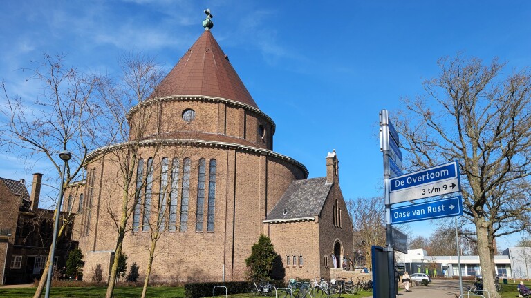 Ronde bakstenen kerk met koepeldak onder een heldere blauwe lucht, met een wegwijzer naar "De Overtoom" en "Oase van Rust".