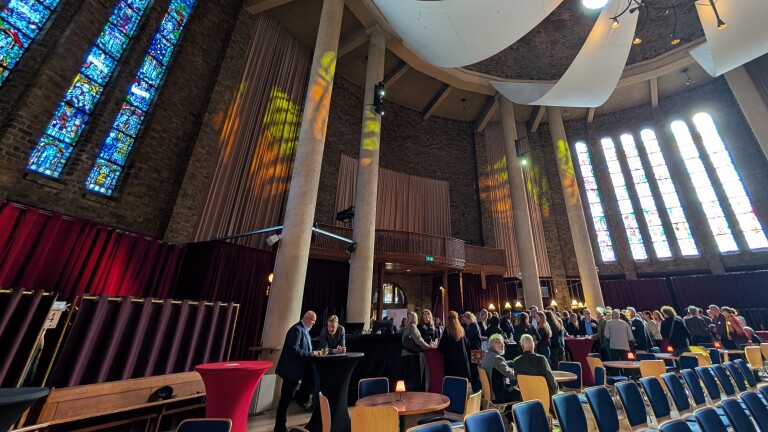 Mensen verzamelen zich in een kerkachtige ruimte met hoge glas-in-loodramen en pilaren, bij meerdere tafelgroepen.