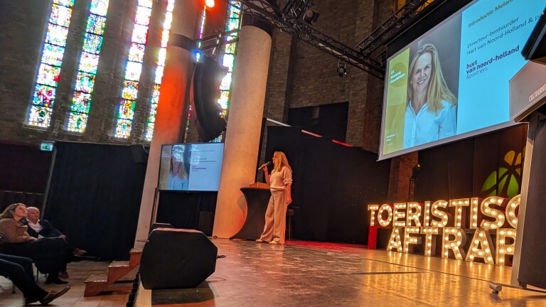 Vrouw op podium geeft presentatie in een kerk met glas-in-loodramen; grote schermen tonen haar afbeelding en naam.