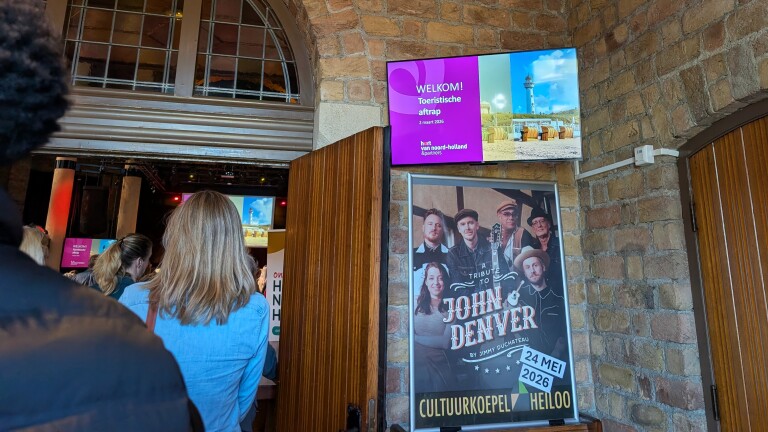 Mensen bij de ingang van een evenement met een scherm dat "Welkom! Toeristische aftrap" toont en een poster voor een John Denver tribute.
