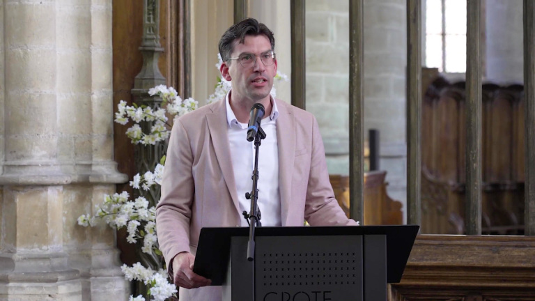 Man in een lichtgekleurde blazer spreekt in een microfoon voor een lessenaar in een sfeervolle kerkelijke omgeving.