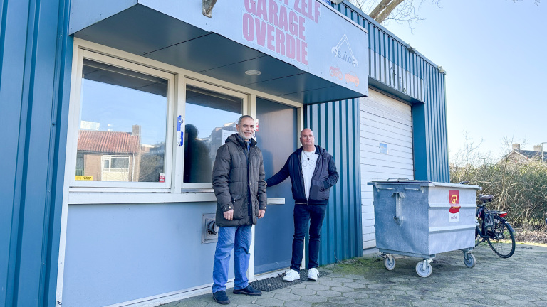 Twee mannen staan voor een garage met het bord "Zelfgarage Overdie". Er is een afvalcontainer en een fiets naast hen.