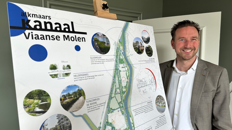 Man in een pak naast een informatiebord over het Alkmaars Kanaal, met afbeeldingen en tekst over verschillende locaties.