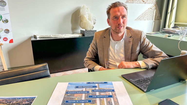 Man in pak zit aan bureau met laptop en papieren met straatnamen; achtergrond bevat kunstwerk en presentatiebord.