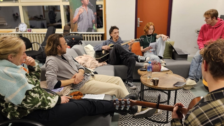 Een groep mensen zit op banken in een informele ruimte; één speelt gitaar, anderen luisteren en lezen notities.