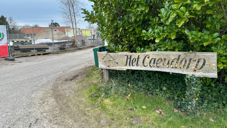 Houten bord met de tekst "Het Caendorp" voor groene struiken, naast een bouwterrein met hekken en stapels materialen.