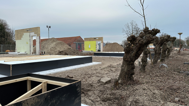 Bouwplaats met gedeeltelijk afgebroken muren en funderingen in voorbereiding, met op de achtergrond stapels aarde en geknotte bomen.