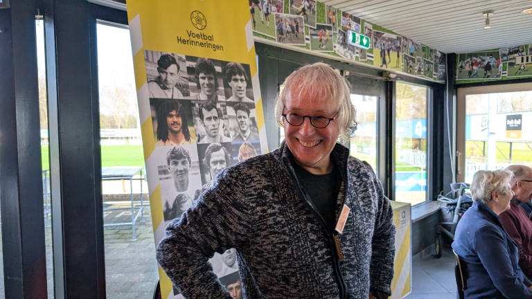 Man met bril en krullend haar lacht voor een poster met oude voetbalfoto's in een clubhuisomgeving.