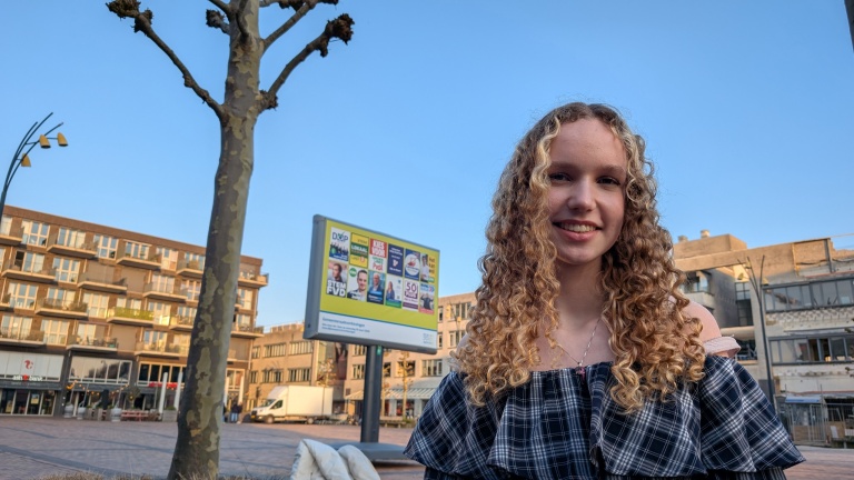 Jongere met krullend haar glimlacht, staand op een plein met een boom en een bord met affiches op de achtergrond.