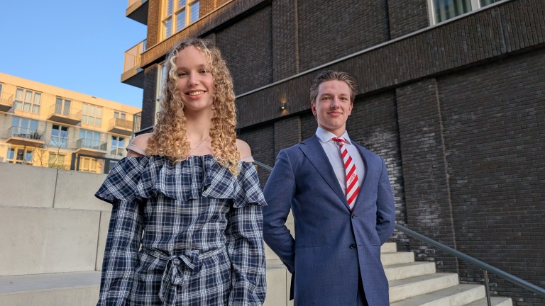 Twee jonge mensen poseren buiten voor een modern gebouw; de vrouw draagt een geruite jurk, de man een pak met een stropdas.