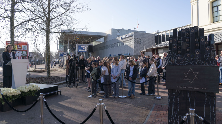 Een groep mensen verzameld bij een herdenkingsmonument met Davidsterren, terwijl een spreker aan een podium een toespraak houdt.