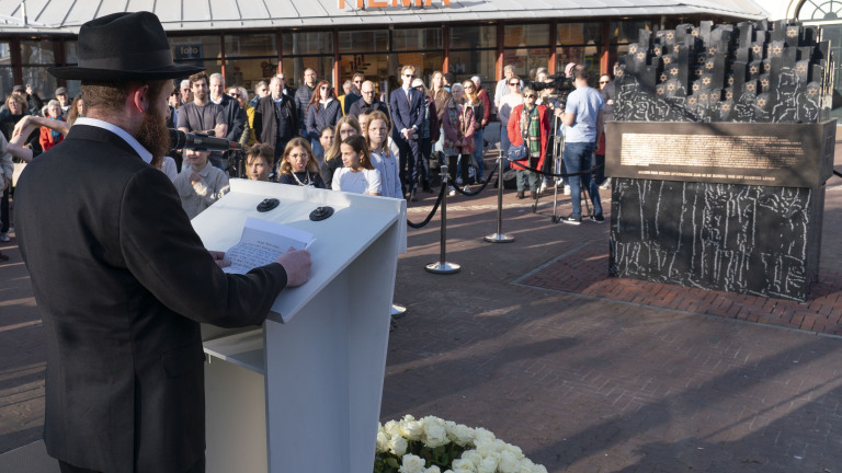 Een man in een zwart pak houdt een toespraak bij een herdenkingsmonument, terwijl een groep mensen toekijkt.