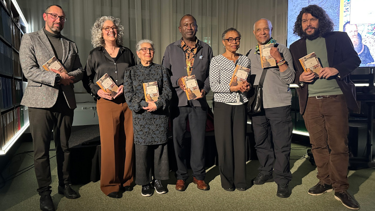 Groep van zeven mensen poseert met boeken in hun handen, staand voor een rij boekenplanken in een verlichte ruimte.