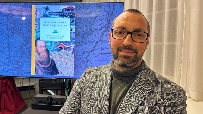 Man in een ruiten colbert voor een scherm met een boekcover getiteld "Alkmaar en het Slavernijverleden".