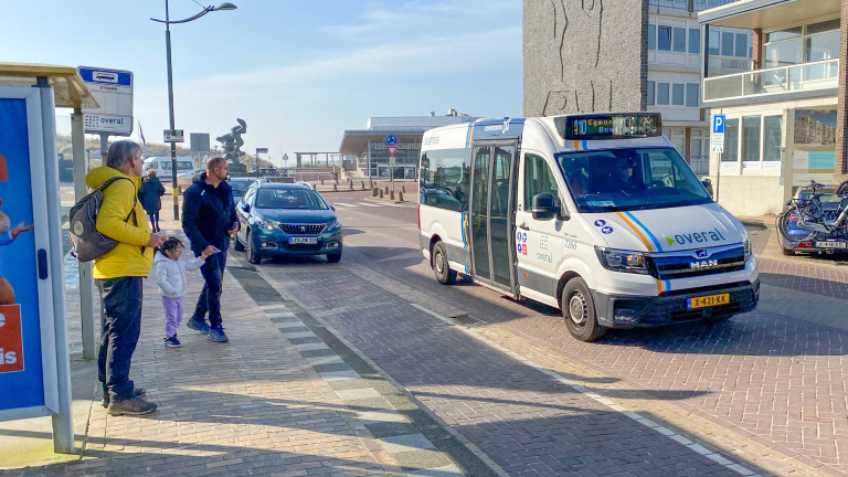 Mensen wachten bij een bushalte terwijl een kleine witte bus nadert met "overal" branding en lijn 410 op de display.
