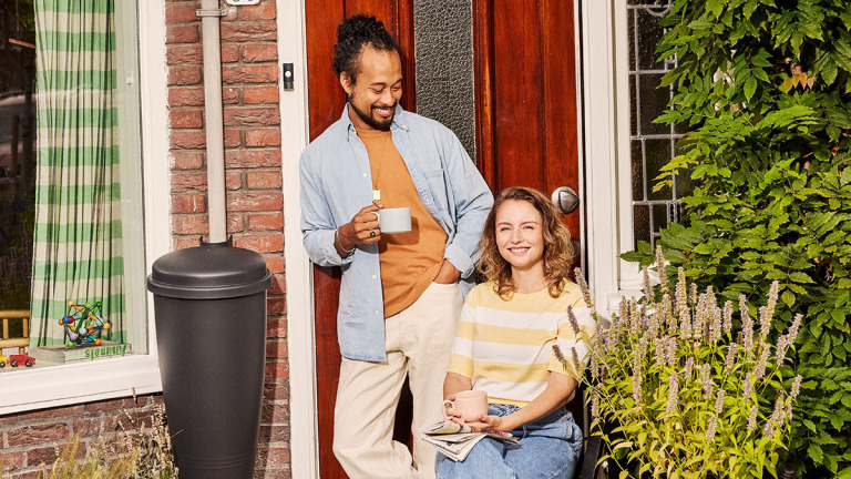 Een man en een vrouw drinken koffie voor een rode deur, omgeven door planten en een bak met groene gordijnen.
