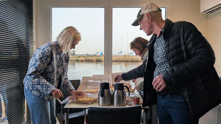 Mensen pakken gebakjes van een tafel in een kamer met glazen deuren, met uitzicht op een waterkant en voorbijrijdende voertuigen.