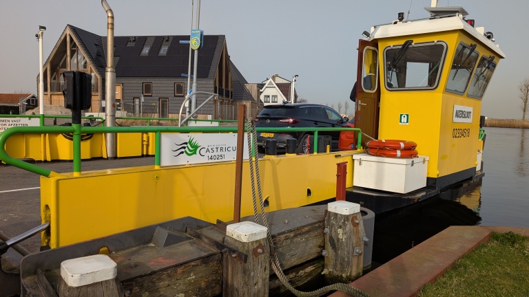 Geel veerbootje genaamd Akersloot met auto aan boord, vastgemaakt aan een pier, op de achtergrond enkele huizen.