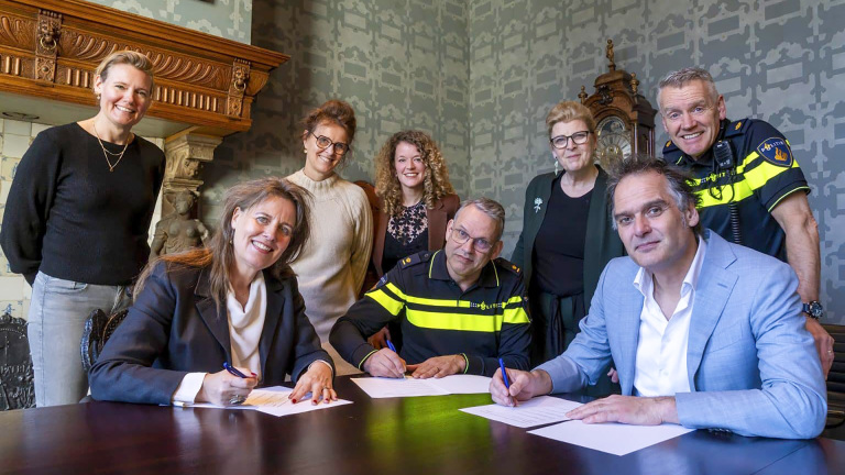 Groep mensen poseert in een kamer met een open haard; drie personen tekenen een document.