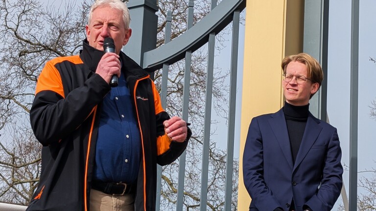Twee mannen staan buiten, de linker man spreekt in een microfoon en draagt een zwart met oranje jas, de rechter man draagt een blauw pak.