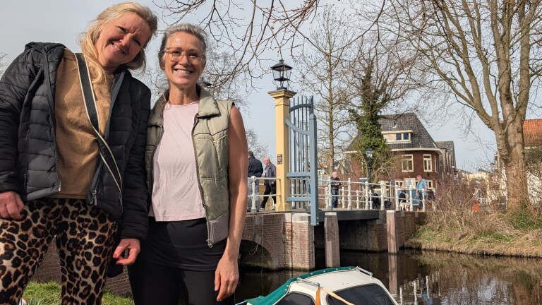 Twee vrouwen staan glimlachend naast een vaart met op de achtergrond een brug en bomen.