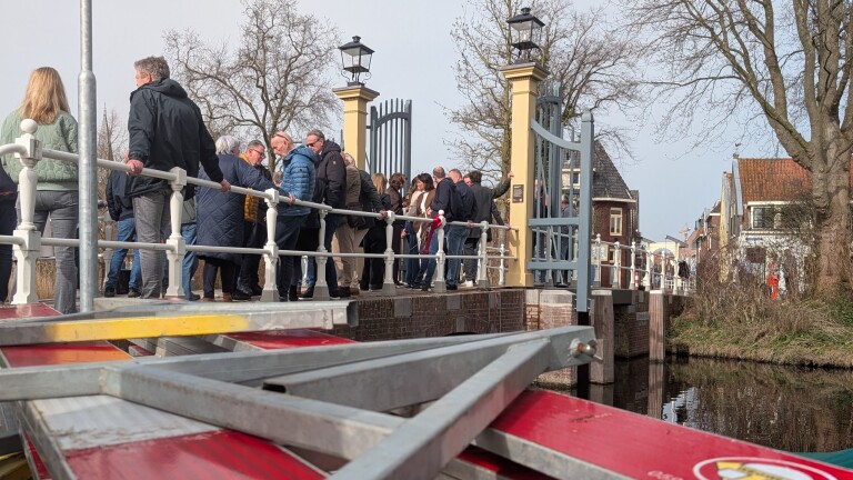 Mensen staan op een brug over een rustige gracht, omgeven door bomen en historische gebouwen.