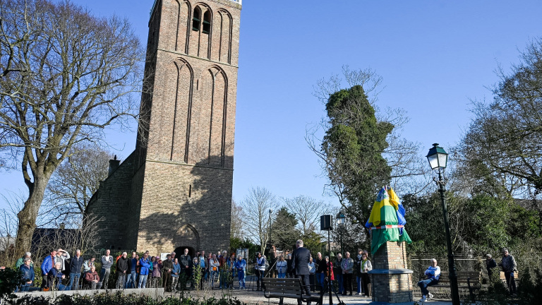 Mensen verzameld voor een oude kerktoren en een met doeken bedekte gedenksteen op een zonnige dag.