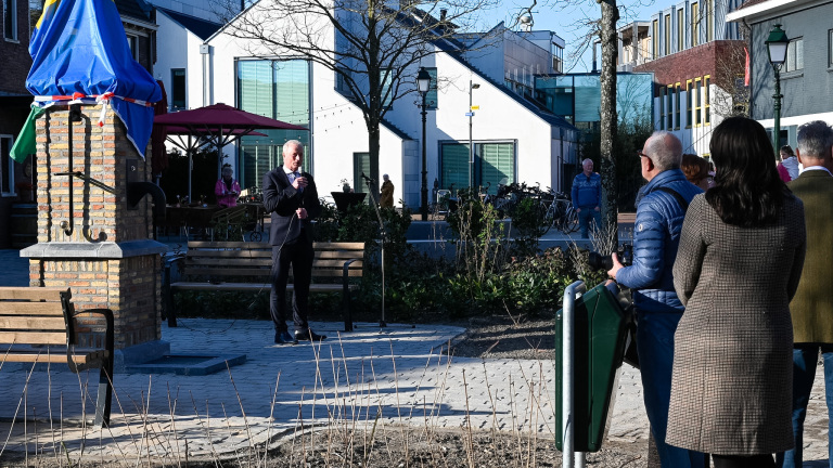 Een man in pak houdt een toespraak naast een bedekt standbeeld, terwijl mensen luisteren op een zonnig plein.