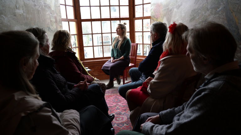 Een groep mensen zit in een kamer met een groot raam, terwijl een vrouw voor hen spreekt.