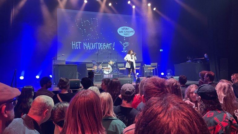 Vrouw in witte jas op podium met microfoon tijdens een evenement genaamd "Het Nachtdebat," omringd door publiek en belichting.